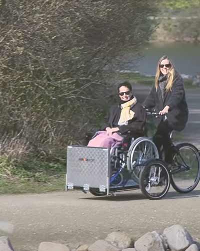 Frau fährt eine andere Frau im Rollstuhl auf einem Lastenfahrrad